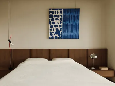 A minimalist bedroom featuring a white bed with a wooden headboard and side tables, adorned with a blue and white abstract pa
