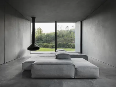 Modern minimalist living room with concrete walls, floor, and furniture, featuring a suspended fireplace and a large window o