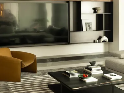 A modern, minimalist living room featuring a wooden wall unit, a leather armchair, a marble coffee table, and a large flat-sc