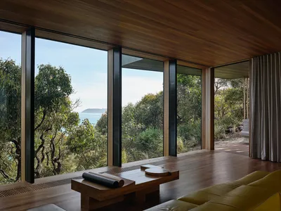 A minimalist bedroom featuring wooden flooring and a glass wall with a view of lush greenery and the ocean.