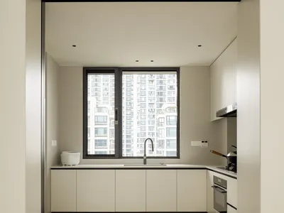 A modern minimalist kitchen with white cabinets and a large window overlooking urban buildings.
