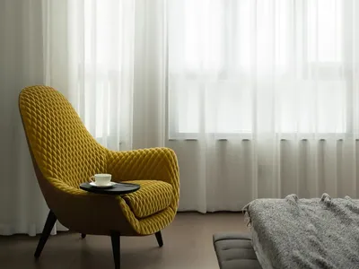 A minimalist bedroom with a yellow velvet armchair, grey bed, and sheer curtains.