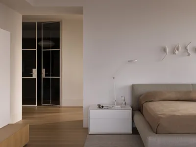 A minimalist bedroom with neutral-toned walls, furniture, and bedding, featuring a simple bed and a white lamp on a white sid