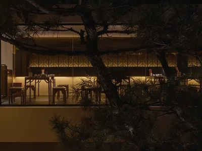 A modern restaurant interior with wooden tables and chairs, illuminated by warm lighting, and featuring a tree in the backgro