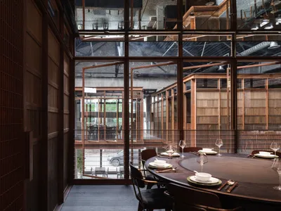 A modern restaurant dining room with a large round table and chairs, made of wood and featuring large windows.