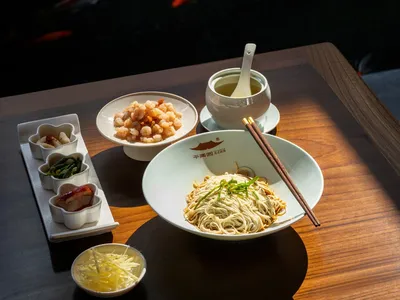 A modern restaurant setting featuring a table with a bowl of noodles, a bowl of shrimp, small dishes of various condiments, a