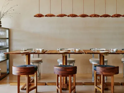Modern restaurant interior with wooden tables and stools, minimalist design, and pendant lighting.