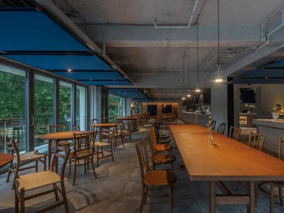 Industrial-style restaurant interior with wooden tables and chairs, concrete ceiling, and large windows.