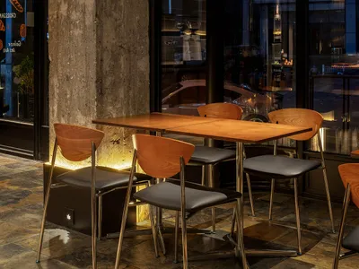 A modern restaurant interior with wooden tables and chairs, glass windows, and a stone wall.