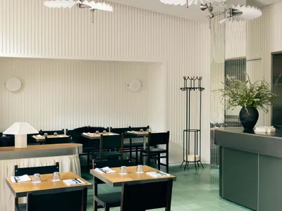 Interior of a minimalist restaurant featuring concrete walls, wooden tables, and black chairs with green plants on the bar co