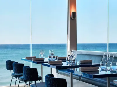A modern restaurant interior featuring tables, chairs, and wine glasses with a glass wall overlooking the ocean.