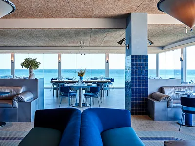 A modern restaurant interior with sea views, featuring blue sofas, tables, chairs, and decorative vases.