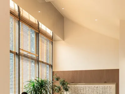 A modern restaurant interior with wooden seating, white cushioned benches, and a large window with blinds allowing natural li