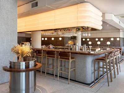A modern restaurant interior featuring a tiled bar with sleek wooden stools and a contemporary light fixture overhead.