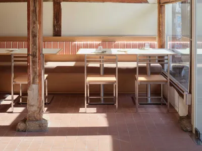 Modern minimalist restaurant interior with wooden seating, metal tables, and terracotta floor tiles.