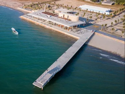 A modern, concrete restaurant building adjacent to a beach with a long jetty extending into the sea.