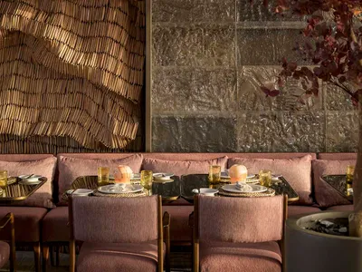 A modern restaurant interior with a woven textile wall and a textured concrete backdrop, featuring brass chairs and tables wi