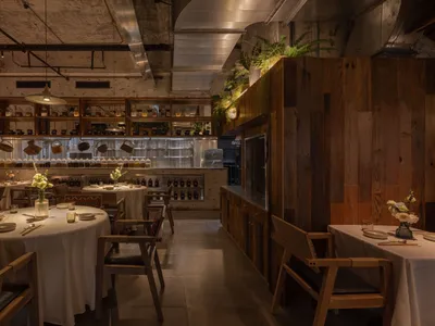 A warm, industrial-themed restaurant interior with wooden furniture, concrete accents, and greenery.