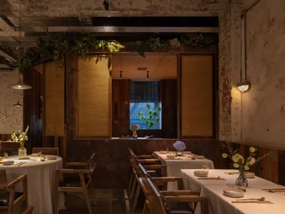 Industrially styled restaurant interior with wooden tables and chairs, white tablecloths, and green plants.