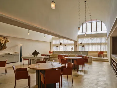 A spacious, modern restaurant interior featuring concrete tables and chairs with high ceilings, pendant lights, and arches.