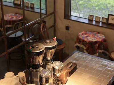 Coffee shop with stainless steel coffee grinders in foreground, tile counter, wooden chairs, and colorful tablecloths.