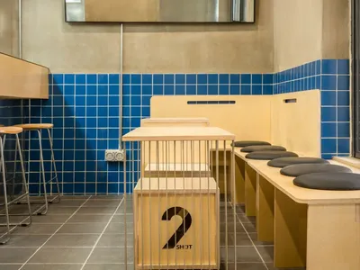A modern industrial-style restaurant interior featuring blue tiled walls, wooden tables, and metal stools.