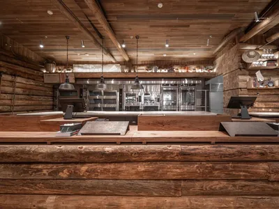 A spacious, rustic restaurant interior with exposed wooden logs on the walls and ceiling, and a modern kitchen area in the ba