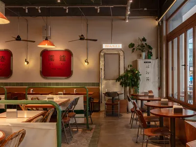 A modern restaurant interior with wooden tables, concrete flooring, metal chairs, and green plants.