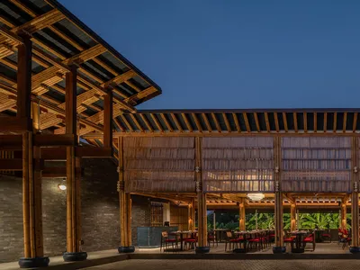 Outdoor area of a modern restaurant featuring bamboo and wooden structures, with tables and chairs arranged under a large roo