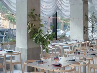Modern minimalist restaurant interior with concrete walls, glass window treatments, and wooden tables and chairs.
