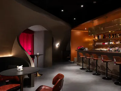 Modern interior design of a restaurant featuring a metal bar counter with leather stools, concrete floor, and glass shelves.