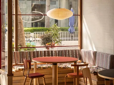 A cozy cafe breakfast area featuring wooden furniture and textile seating, with large windows showcasing outdoor greenery and