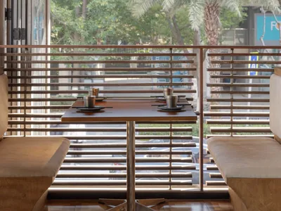 Cafe interior with wooden tables and chairs, large windows showing palm trees, and exposed brick wall