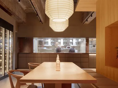 Modern restaurant interior featuring a dining area with wooden tables and chairs, concrete walls, and pendant lighting.