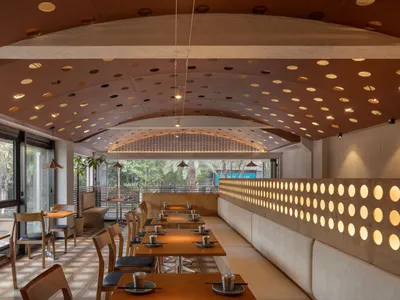 A modern restaurant interior featuring wooden tables, fabric seating, and perforated metal accents.