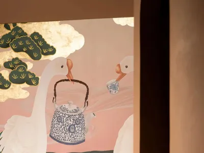 一張描繪咖啡廳室內的圖像，粉金色壁畫上繪有手持茶壺與茶杯的天鵝，背景為靠枕