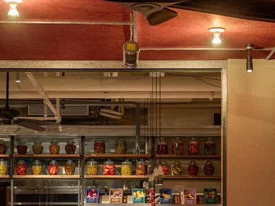 Industrial-style restaurant bar area with wooden tables, metal chairs, glass jars on shelves, and fabric curtains.