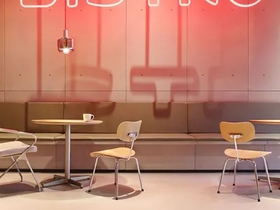 A minimalist industrial-style restaurant interior with exposed concrete walls, a red neon sign reading 'BISTRO', and simple s