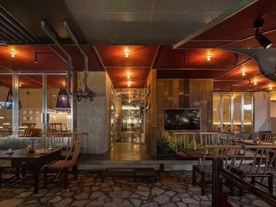 An industrial-style restaurant interior with concrete walls, wooden tables and chairs, and glass ceiling panels.