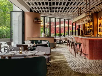 A modern restaurant bar interior featuring wooden furniture, glass shelves, metal stools, and fabric accents.