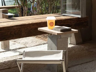 A modern coffee shop exterior with wooden seating and concrete structures, surrounded by greenery, with a cup of drink on a s