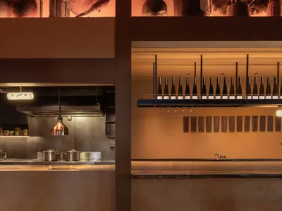 A modern, industrial restaurant bar with concrete, metal, and wood materials, featuring a wine bottle display and overhead li