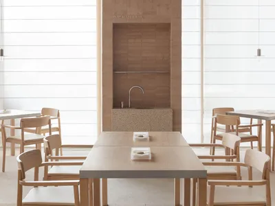 A modern restaurant interior with minimalist design, featuring wooden tables, stone walls, and large windows.