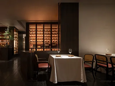 An elegant, dimly lit dining room with a well-lit wine collection in the background and a single table set for a meal.