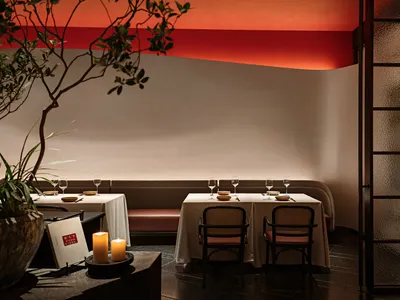 A cozy modern restaurant interior with white walls, red ceiling accents, fabric seating, candles, and potted plants.