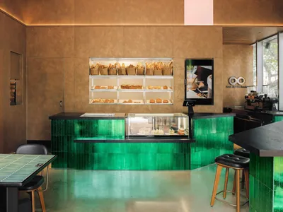 A coffee shop with a green tile and black counter displaying pastries and cakes in a glass display case.