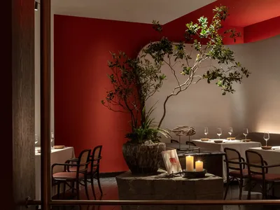 A modern restaurant interior with red walls and a potted plant, tables set with white tablecloth, candles, and glasses.