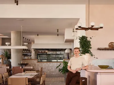 A man wearing a beige shirt and brown pants is seated at the bar of a modern restaurant, with clean lines and a minimalist ae