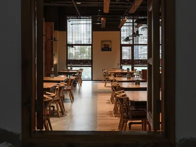 Industrial-style restaurant interior with wooden tables and chairs, large windows, and exposed beams.