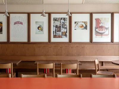 A modern restaurant interior featuring wooden tables, chairs, and benches with framed artworks on the wall.